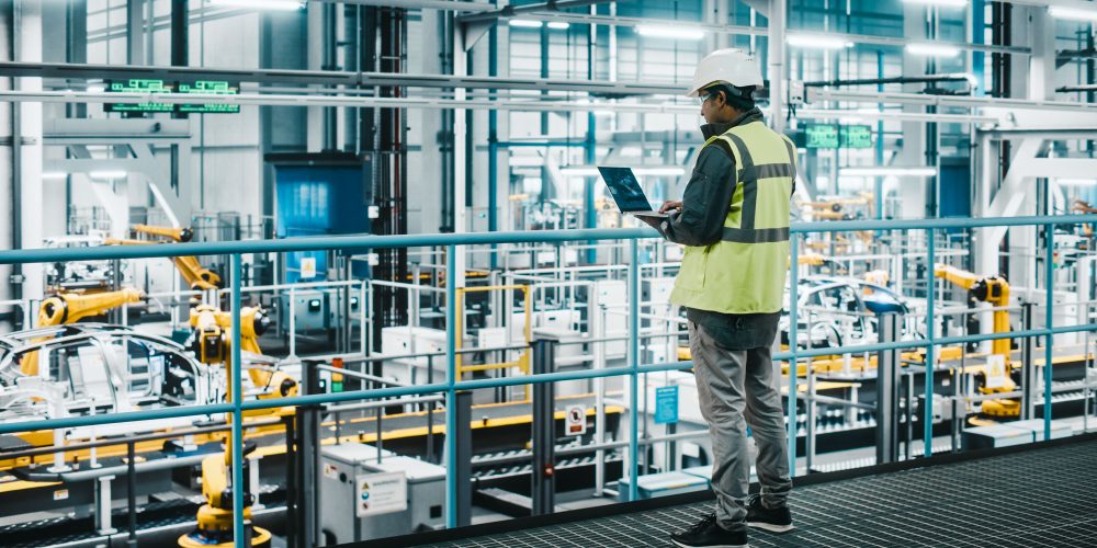 Car Factory Engineer in High Visibility Vest Using Laptop Computer. Automotive Industrial Manufacturing Facility Working on Vehicle Production with Robotic Arms Technology. Automated Assembly Plant.